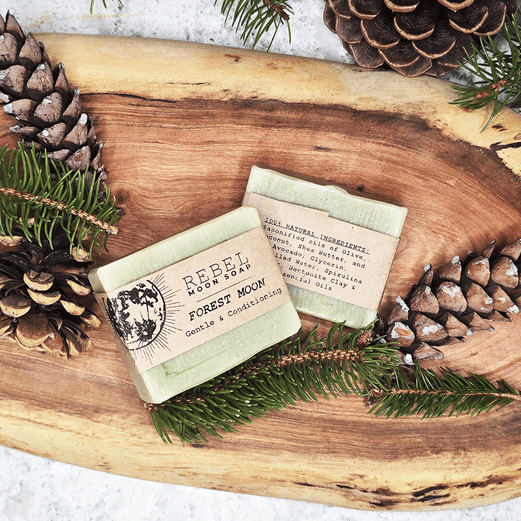Two bars of soap with 'Rebel Moon Soap' branding on a wooden surface with pine branches and pinecones.